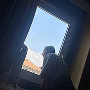 Fiona participe au concours pour gagner de l'argent avec cette photo : person, cat, window, curtain, sky, clouds, indoor, windowsill, standing, leaning, silhouette, daylight, building, roof, profile, casual_clothing, side_view, pet, curious, calm