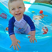 Nathaël a rejoint le concours — aidez-le/la à gagner de superbes lots ! toddler, child, smiling, swimsuit, pool, inflatable_pool, water, outdoor, summer, playful, happy, baby, grass, sunlight, toy_ball, blue, cute, fun, face, hands
