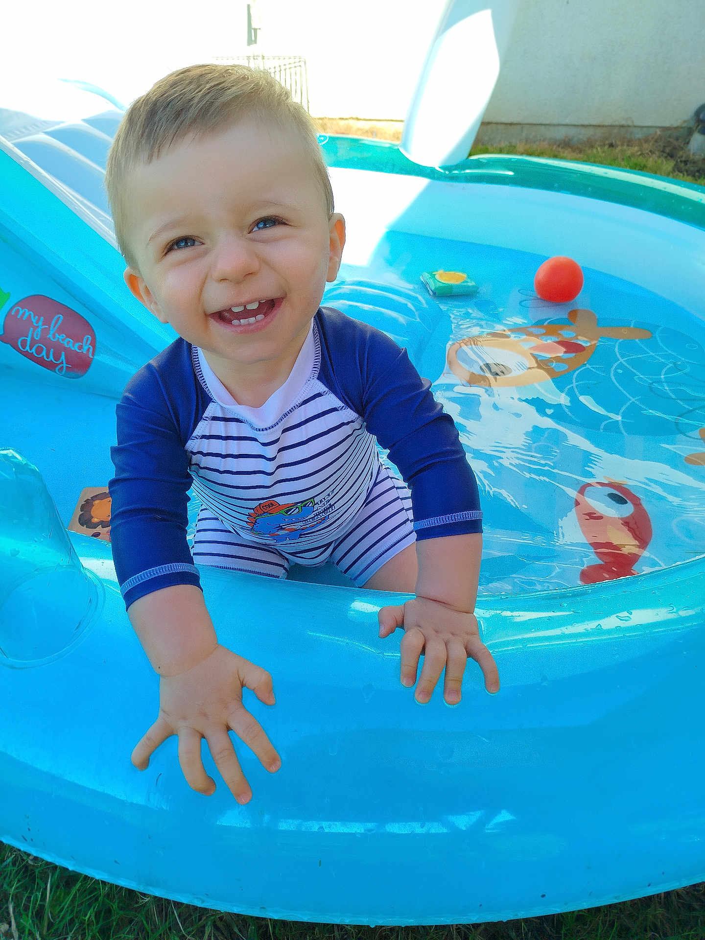 Nathaël a rejoint le concours — aidez-le/la à gagner de superbes lots ! toddler, child, smiling, swimsuit, pool, inflatable_pool, water, outdoor, summer, playful, happy, baby, grass, sunlight, toy_ball, blue, cute, fun, face, hands