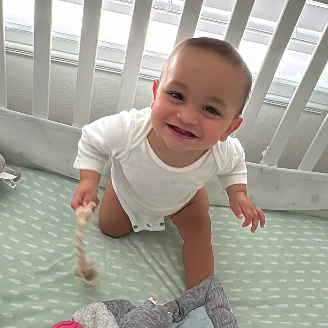 Derian joined the competition — help win amazing prizes! baby, crib, toy, blanket, smile, child, infant, white_onesie, nursery, happy, indoors, cute, playful, face, person, soft_toy, bed, room, young_child, crawling