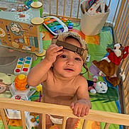 Derian is registered to the contest to win money with this photo: toddler, child, playpen, toy, cap, barefoot, indoor, colorful, playmat, stuffed_animal, basket, wooden_bars, curious, baby, diaper, smiling, standing, furniture, person, home