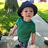 toddler, child, hat, green_shirt, plaid_shorts, shoes, outdoor, sidewalk, tree, grass, sunlight, daytime, curious, portrait, person, young_child, casual_clothing, nature, summer, background