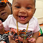 Kavari is registered to the contest to win money with this photo: adult_hand, baby, cap, child, closeup, clothing, cute, expression, face, gold_chain, happy, holding, indoor, jewelry, orange_bow, person, portrait, smile, tattoo, white_shirt
