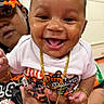 adult_hand, baby, cap, child, closeup, clothing, cute, expression, face, gold_chain, happy, holding, indoor, jewelry, orange_bow, person, portrait, smile, tattoo, white_shirt