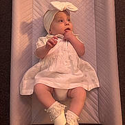 Mýa-Rae is registered to the contest to win money with this photo: baby, child, infant, white_dress, headband, socks, shoes, changing_pad, quilted, carpet, cute, lying_down, indoors, portrait, adorable, curious, hand, finger, soft_lighting, young_child