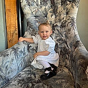 Olive is registered to the contest to win money with this photo: toddler, child, white_dress, black_shoes, armchair, floral_pattern, smile, hair_buns, indoor, furniture, cute, portrait, sitting, young_child, person, happy, cozy, home, small_child, fashion