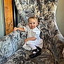 toddler, child, white_dress, black_shoes, armchair, floral_pattern, smile, hair_buns, indoor, furniture, cute, portrait, sitting, young_child, person, happy, cozy, home, small_child, fashion