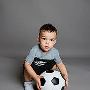 Dylan is registered to the contest to win money with this photo: toddler, child, soccer_ball, gray_background, nike, shorts, sneakers, curious, crouching, studio, indoor, sport, toy, footwear, clothing, portrait, looking_up, young_child, casual, play