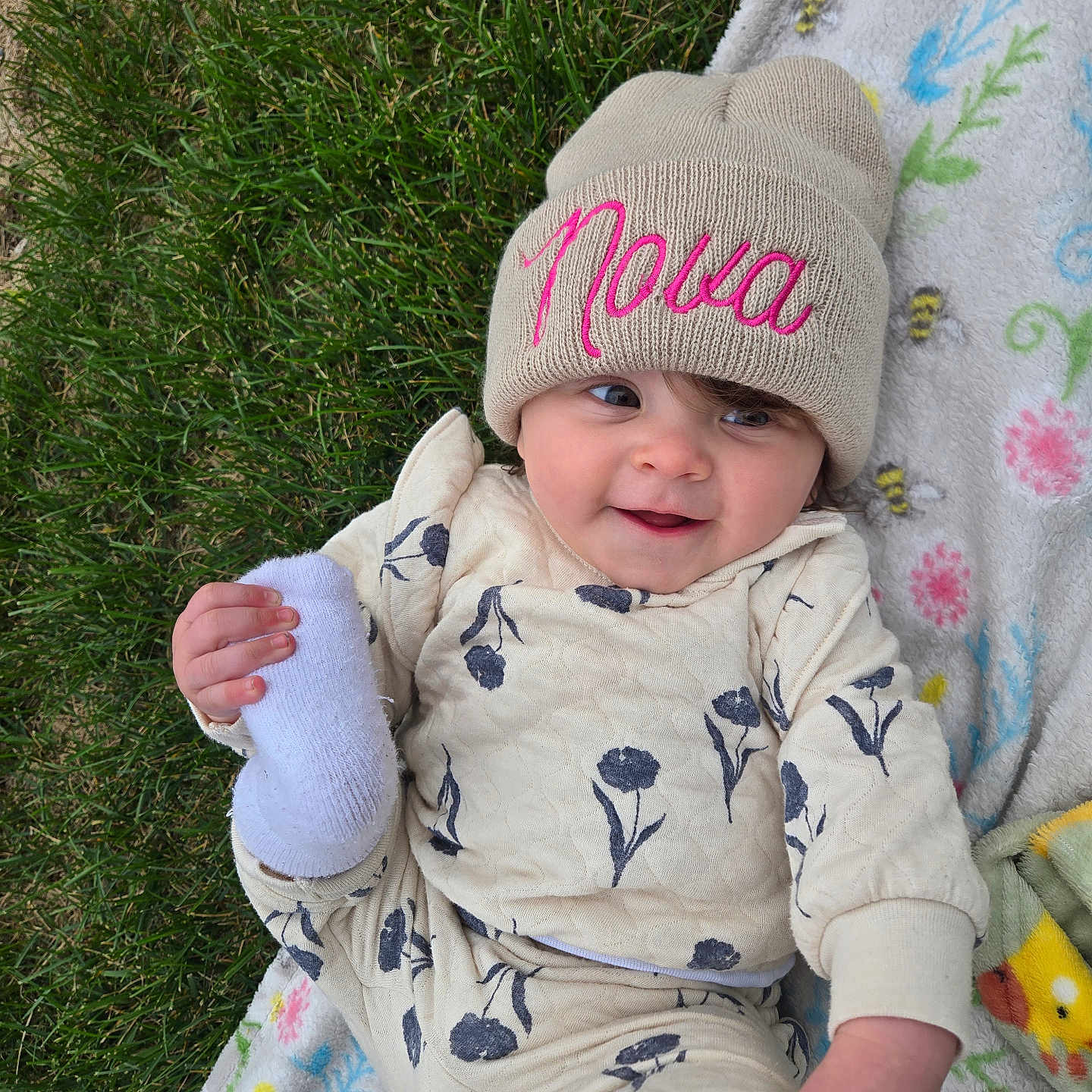 Nova is registered to the contest to win money with this photo: baby, beanie, blanket, child, clothing, cute, floral_pattern, foot, grass, hand, happy, hat, infant, nature, outdoor, person, playful, portrait, smiling, toddler
