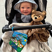 Koby is registered to the contest to win money with this photo: baby, child, stroller, hooded_jacket, blanket, teddy_bear, soft_toy, book, farm_animals, indoor, floor_tiles, smiling, happy, cute, person, infant, seated, cozy, playful, comfort