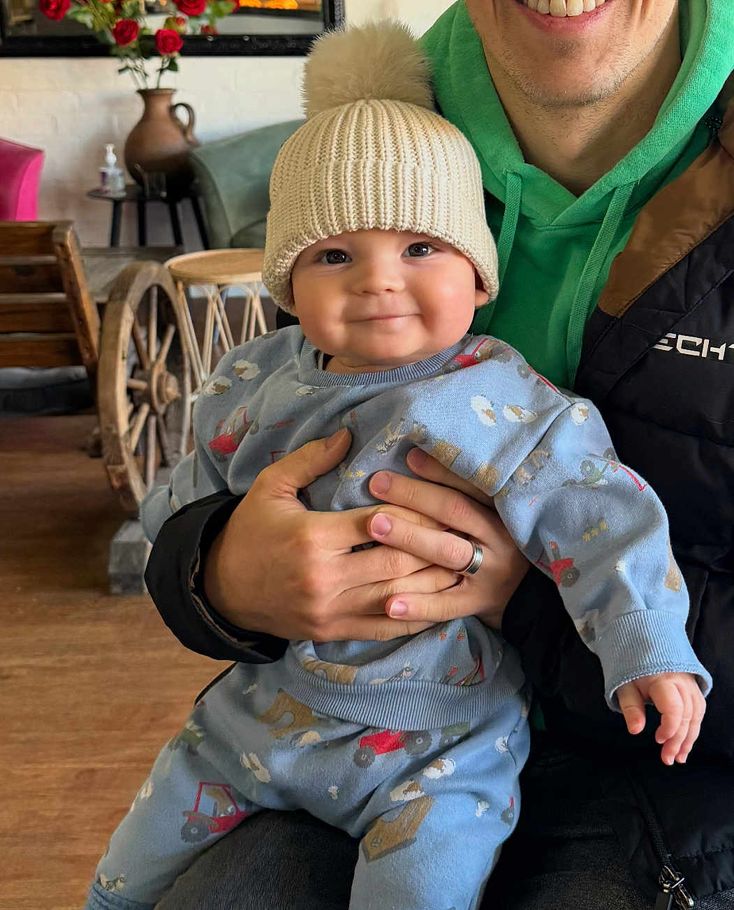 Koby joined the competition — help win amazing prizes! baby, child, person, hat, knit_hat, sweater, blue_clothing, smile, hand, adult, green_hoodie, black_jacket, indoor, wooden_furniture, vase, flowers, roses, rustic, cozy, portrait