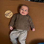 Miller is registered to the contest to win money with this photo: baby, infant, smiling, sweater, pants, socks, blanket, wooden_sign, three_months, lying_down, indoors, cute, child, portrait, cozy, happy, face, person, young_child, clothing