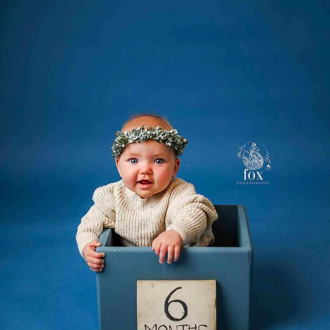 Juniper is registered to the contest to win money with this photo: accessories, baby, bonnet, box, cap, clothing, costume, dress, face, happy, hat, head, jewelry, necklace, newborn, person, photography, portrait, sitting, smile