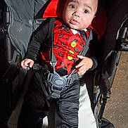 Andre is registered to the contest to win money with this photo: baby, child, stroller, costume, vampire, cape, red, black, straps, seat, wide_eyes, curious, infant, person, cute, portrait, indoors, clothing, baby_seat, expression
