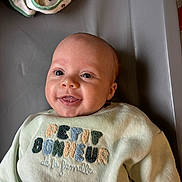 Théo a rejoint le concours — aidez-le/la à gagner de superbes lots ! baby, infant, smiling, face, eyes, tongue, sweater, clothing, embroidery, text, fabric, soft, lying, indoor, cute, child, person, portrait, head, happy