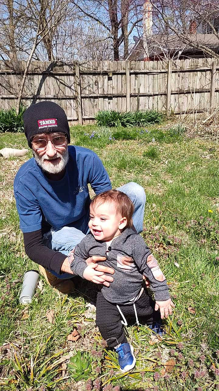 Jason is registered to the contest to win money with this photo: baseball_cap, beard, cap, child, fence, garden, grass, grassland, happy, hat, headwear, landscape, lawn, people_in_nature, person, plant, sitting, smile, soil, spring