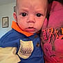 Jasper is registered to the contest to win money with this photo: baby, child, person, indoor, clothing, blue_shirt, orange_collar, face, curly_hair, portrait, expression, furniture, wall_art, sitting, closeup, young_child, skin, head, eyes, holding