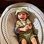 baby, smiling, green_hat, green_outfit, baby_seat, cushion, infant, happy, indoor, wooden_furniture, portrait, child, clothing, cute, sitting, expression, face, small_hands, legs, comfortable
