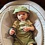 Jasper is registered to the contest to win money with this photo: baby, infant, green_clothing, bucket_hat, baby_seat, cushion, face, hand, legs, wooden_furniture, indoor, portrait, cute, young_child, comfortable, casual, overalls, wristband, looking_at_camera, smile