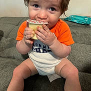 Zayden is registered to the contest to win money with this photo: toddler, child, pudding, food, orange_shirt, white_onesie, couch, furniture, indoor, sitting, hands, face, legs, cute, snack, expression, eyes, hair, person, home