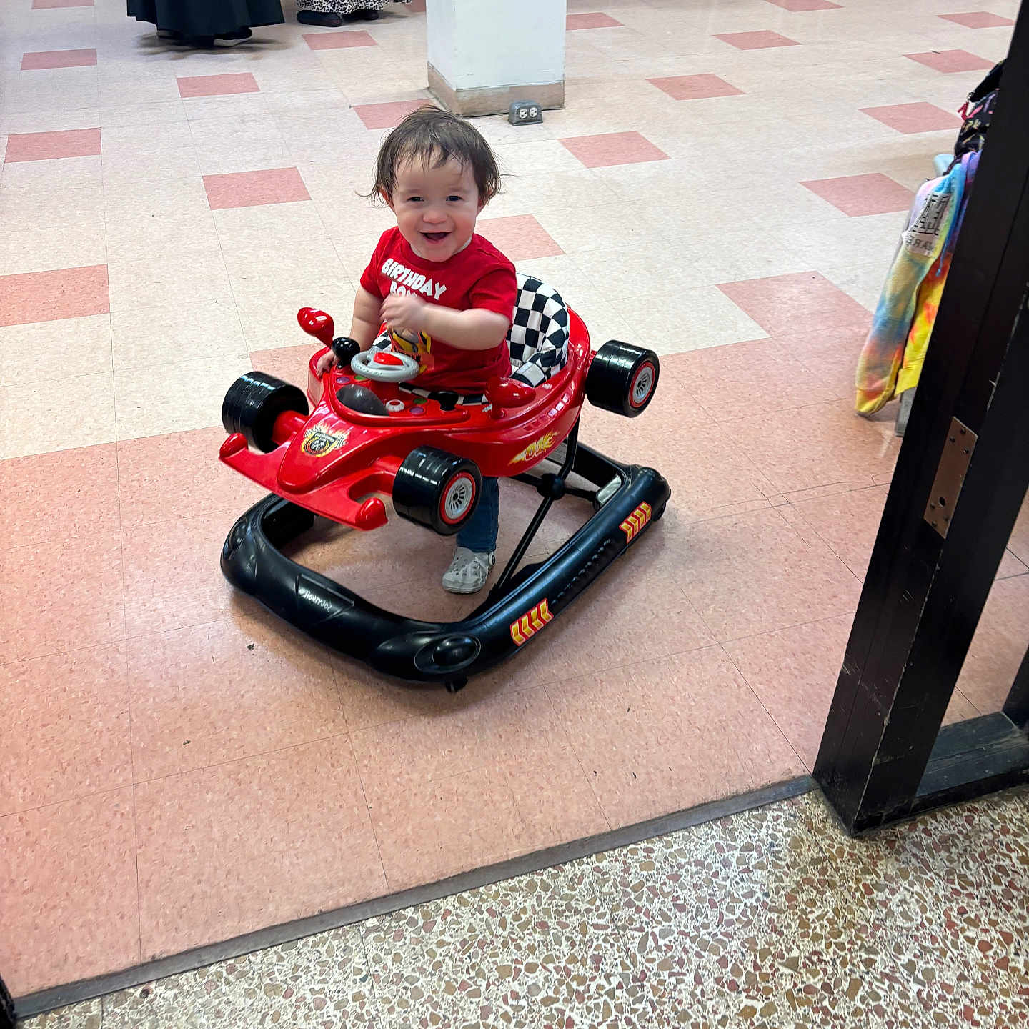Zayden is registered to the contest to win money with this photo: adult, background, birthday, chair, child, clothing, floor_tiles, fun, happy, indoor, person, pillar, play, red_shirt, shoes, smiling, table, toddler, toy_car, walker