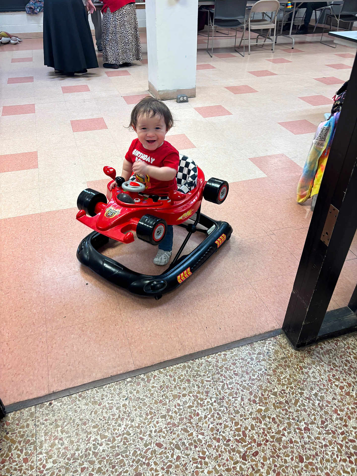 Zayden is registered to the contest to win money with this photo: toddler, child, toy_car, walker, red_shirt, birthday, indoor, floor_tiles, smiling, happy, person, adult, clothing, shoes, table, chair, pillar, background, play, fun