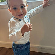 Kais participe au concours pour gagner de l'argent avec cette photo : toddler, child, smiling, standing, indoor, window, hand, long_sleeve_shirt, blue_pants, barefoot, wooden_floor, natural_light, greenery, face, person, happy, cute, casual_clothing, portrait, home