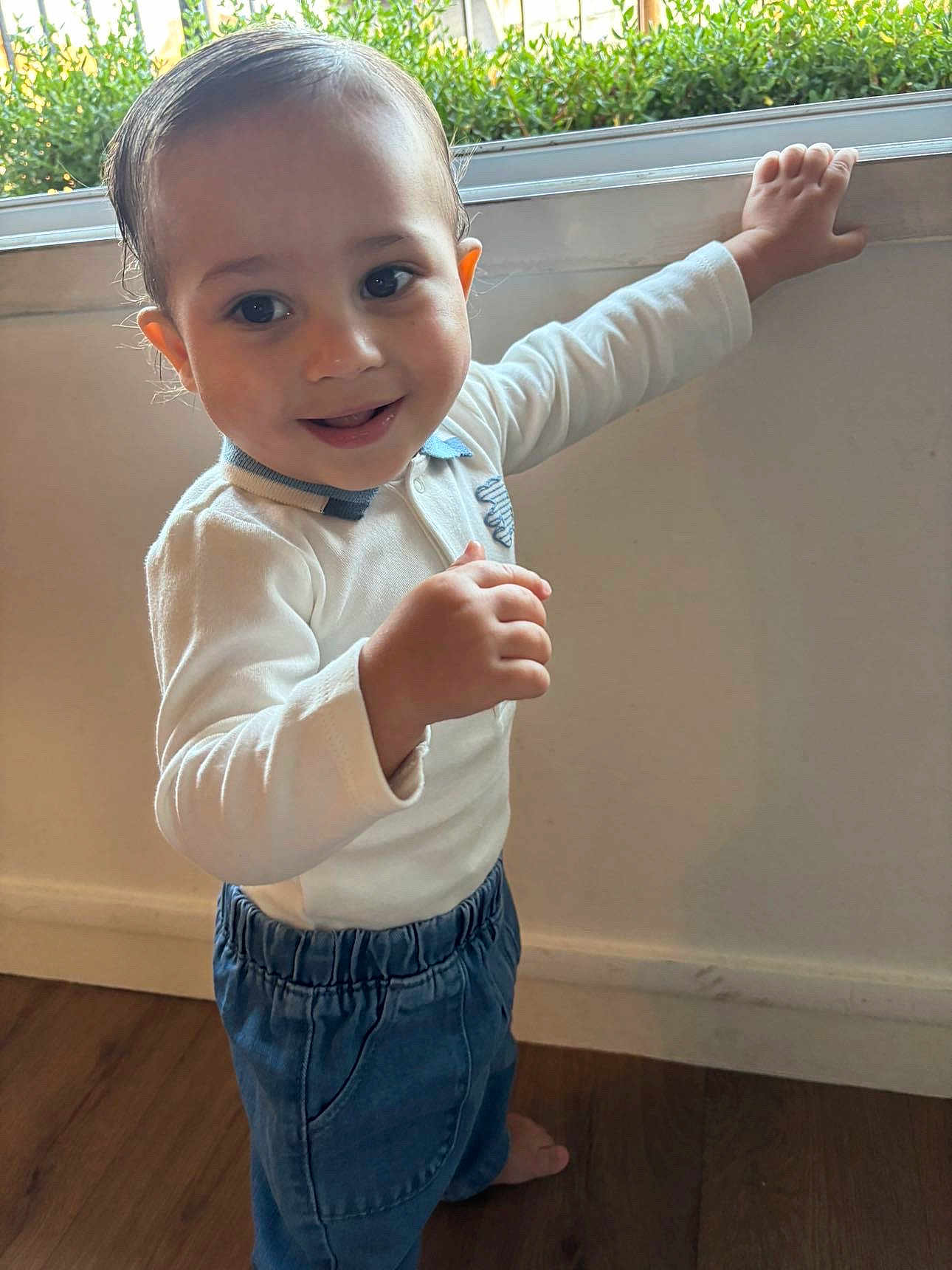 Kais participe au concours pour gagner de l'argent avec cette photo : toddler, child, smiling, standing, indoor, window, hand, long_sleeve_shirt, blue_pants, barefoot, wooden_floor, natural_light, greenery, face, person, happy, cute, casual_clothing, portrait, home