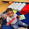 Tamia is registered to the contest to win money with this photo: child, toddler, braided_hair, beads, playing, red_sticks, blue_carpet, cushions, book, jeans, heart_patch, indoor, smiling, happy, floor, casual_clothing, toy, colorful, fun, preschool