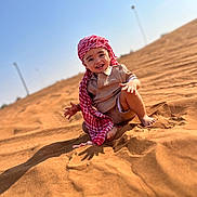 Kyllian participe au concours pour gagner de l'argent avec cette photo : child, toddler, smiling, sand, desert, sunny, blue_sky, headwear, red_scarf, barefoot, outdoor, happy, portrait, clothing, shadow, nature, daylight, cute, young, sitting