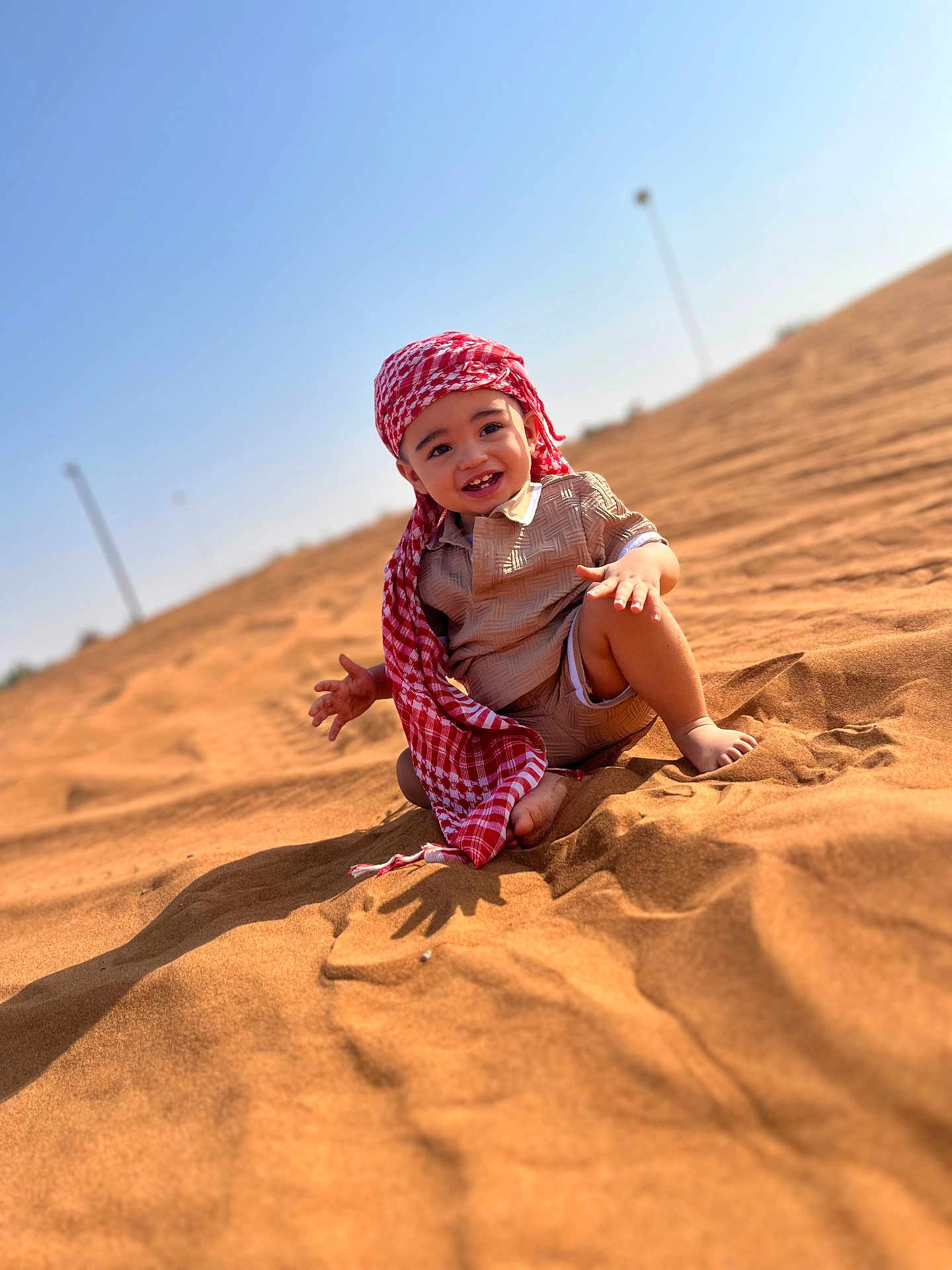 Kyllian participe au concours pour gagner de l'argent avec cette photo : child, toddler, smiling, sand, desert, sunny, blue_sky, headwear, red_scarf, barefoot, outdoor, happy, portrait, clothing, shadow, nature, daylight, cute, young, sitting