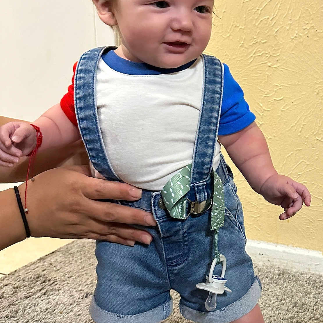 Manuel Maturino Jr is registered to the contest to win money with this photo: blue_shirt, carpet, child, clothing, cute, denim, footwear, hands, indoor, infant, pacifier, person, red_shirt, shoes, shorts, standing, suspenders, textured_wall, toddler, wall