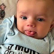 Rylan is registered to the contest to win money with this photo: baby, bed, blanket, blue_clothing, child, close_up, cozy, curious, eyes, face, family, hand, indoors, infant, lying_down, patterned_fabric, photography, portrait, smiling, tongue