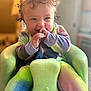 baby, baby_seat, child, closeup, colorful_clothing, curly_hair, cute, face, green_seat, hands, happy, indoor, infant, onesie, playful, seated, smiling, soft_lighting, tie_dye, tongue_out
