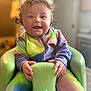 baby, bright_eyes, child, colorful_clothing, cozy, curly_hair, cute, foam_chair, hand_holding, happy, indoors, onesie, person, portrait, seated, smiling, tie_dye, warm_lighting, wet_hair, young_child