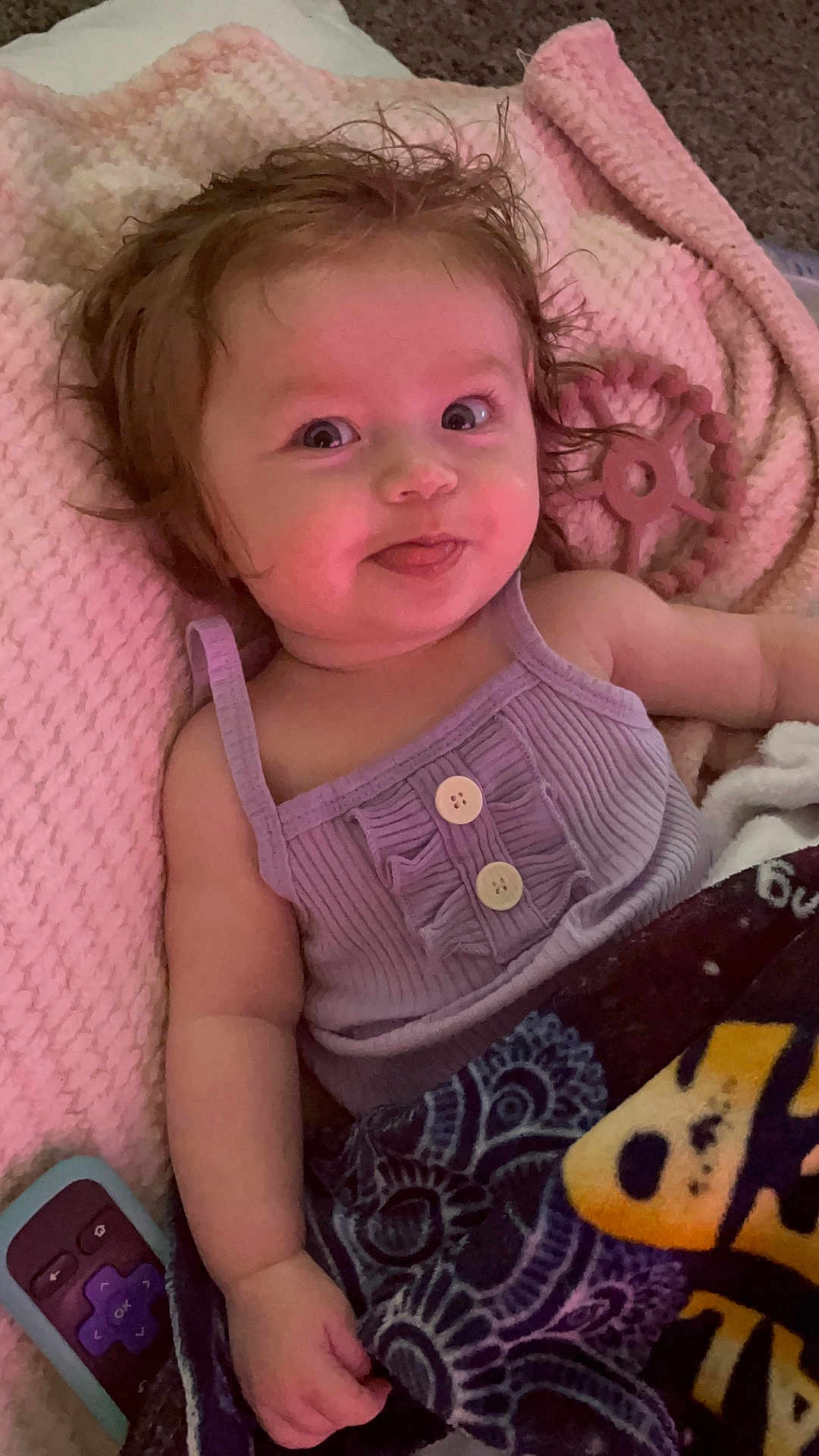 Lilith joined the competition — help win amazing prizes! baby, child, tongue_out, purple_top, blanket, pink_blanket, remote_control, cute, smiling, indoor, toddler, soft_texture, buttoned_top, lying_down, playful_expression, face, arm, hand, hair, cozy