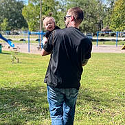 Lilith is registered to the contest to win money with this photo: man, baby, child, park, grass, trees, blue_sky, sunlight, playground, casual_clothing, jeans, black_shirt, sunglasses, smiling, outdoor, holding, walking, nature, daytime, happy