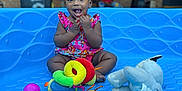 Emmery is registered to the contest to win money with this photo: toddler, child, smiling, sitting, colorful_clothing, plush_toys, ball, blue_surface, outdoor, brick_wall, person_in_background, happy, playtime, cute, baby, toy, casual, daylight, blurred_background, portrait