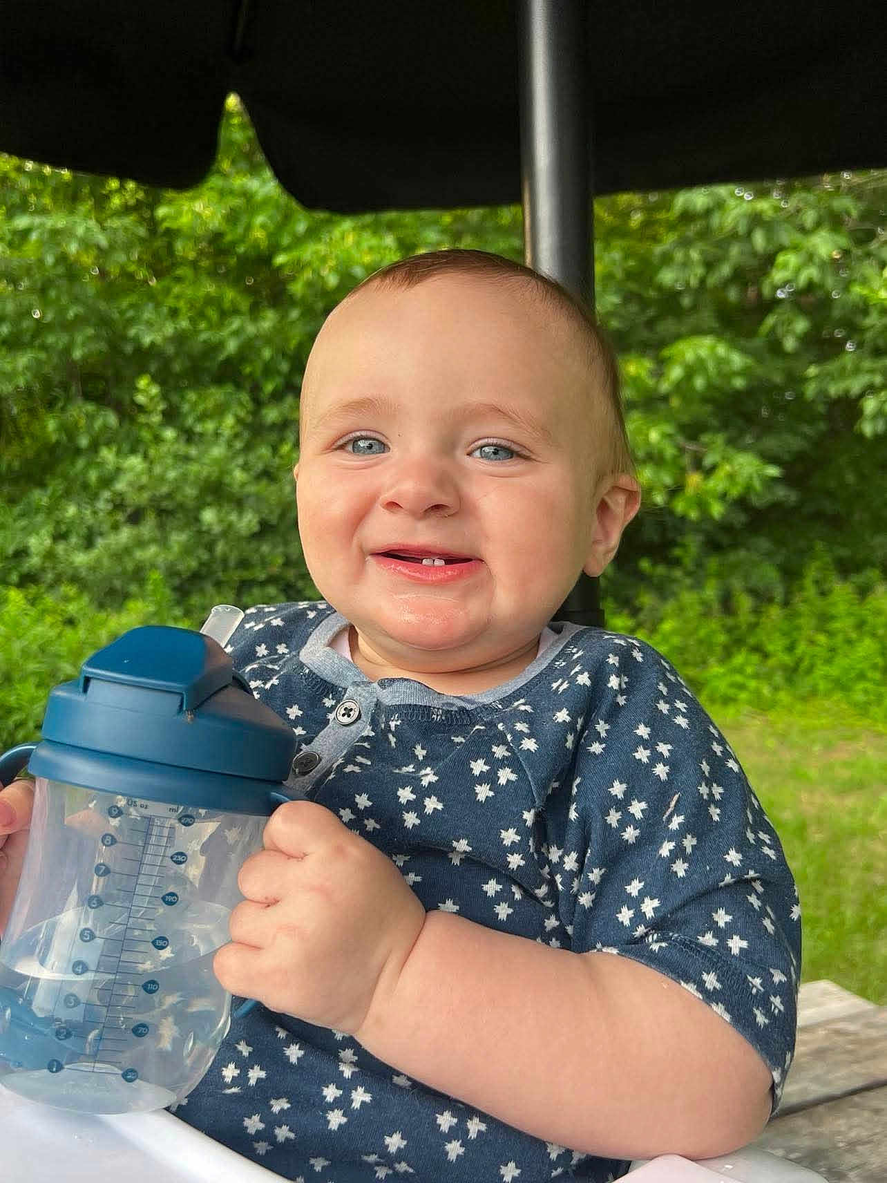 Wilder is registered to the contest to win money with this photo: baby, child, smiling, blue_eyes, sippy_cup, outdoor, greenery, umbrella, happy, tooth, clothing, short_sleeves, nature, table, person, infant, cute, face, hand, daylight