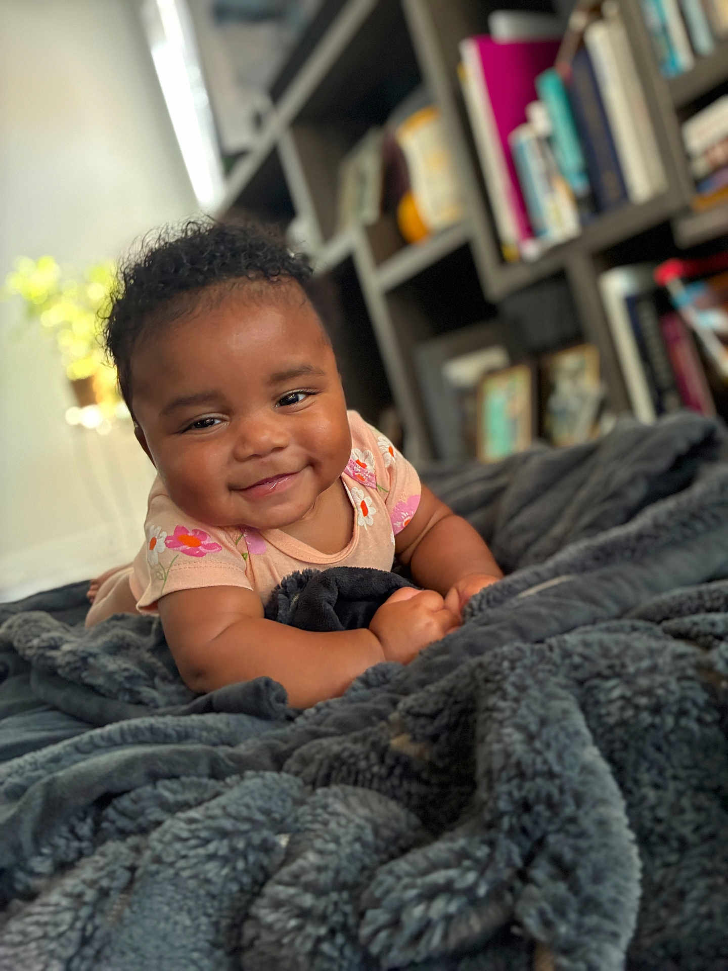 Unique is registered to the contest to win money with this photo: baby, smiling, infant, blanket, curly_hair, onesie, floral_pattern, indoors, bookshelf, books, cozy, soft_light, portrait, cute, child, happy, lying_down, furniture, home, warmth