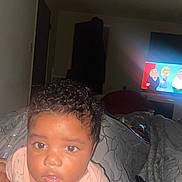 Unique joined the competition — help win amazing prizes! baby, child, curly_hair, indoor, couch, blanket, remote_control, television, cartoon, face, person, clothing, pattern, room, light, young_child, expression, sitting, home, curious