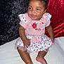 baby, child, infant, smiling, pink_clothing, dress, sitting, barefoot, curly_hair, face, portrait, cute, indoor, fabric, velvet, red_background, black_background, floral_pattern, ladybug, bow