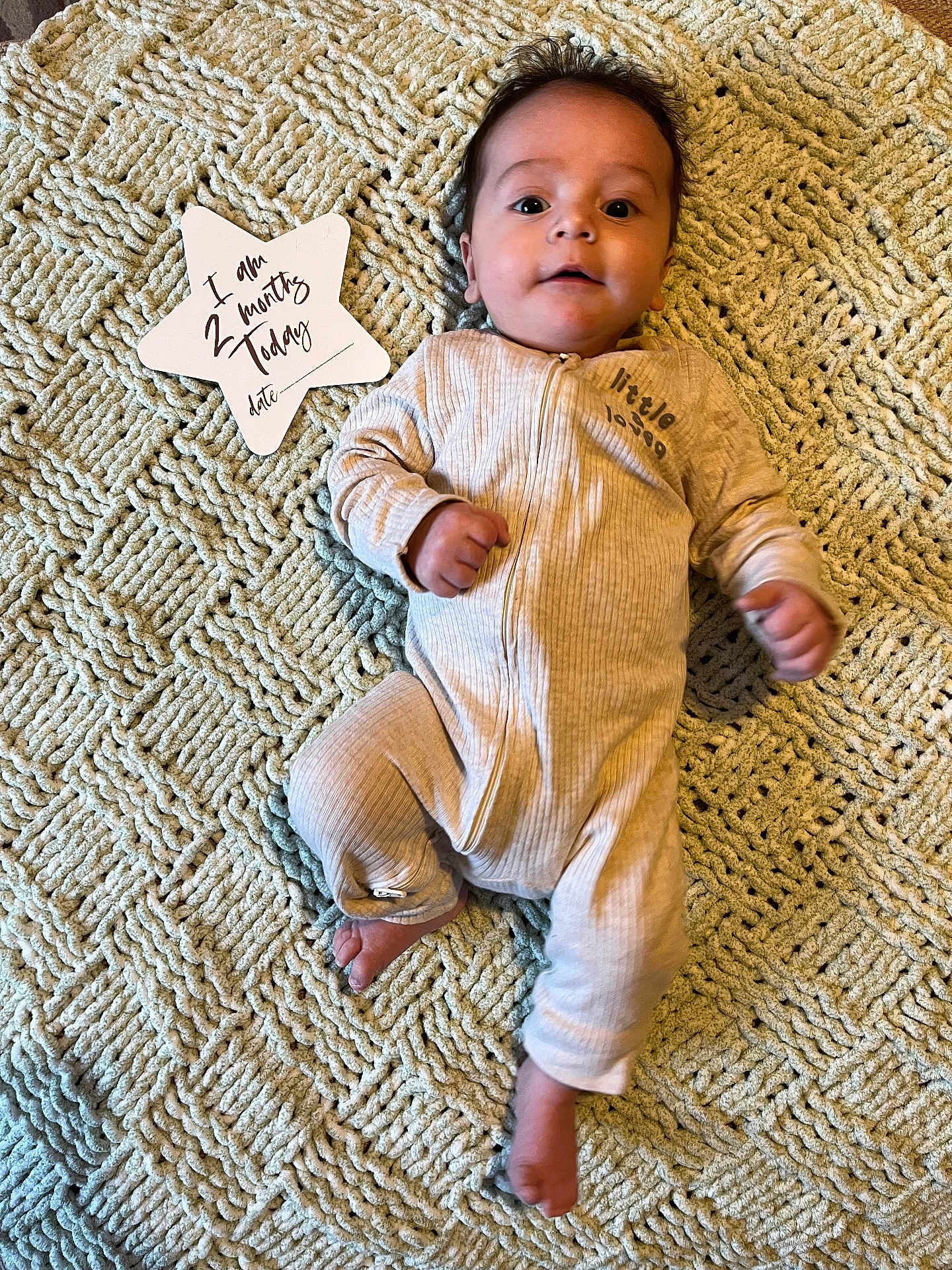 Milo is registered to the contest to win money with this photo: baby, infant, blanket, onesie, star_shaped_sign, milestone, lying_down, indoor, cute, portrait, face, hands, feet, text, milestone_marker, cozy, soft_texture, expression, young_child, newborn