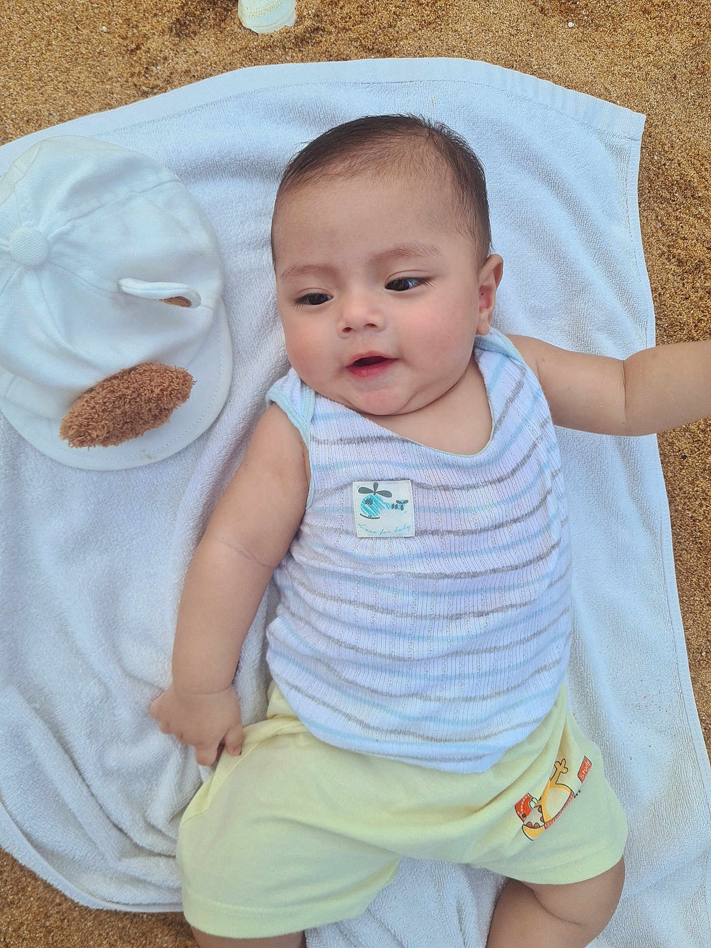 Halley a rejoint le concours — aidez-le/la à gagner de superbes lots ! baby, infant, towel, sand, cap, clothing, striped_top, shorts, outdoor, person, cute, smiling, face, head, skin, young_child, summer, relaxing, lying_down, casual