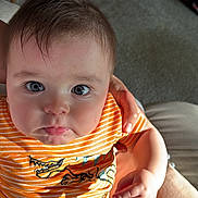 Gael Máximo Pereira is registered to the contest to win money with this photo: baby, child, infant, orange_striped_shirt, dinosaur_print, striped_pants, hand, person, face, eyes, hair, skin, sitting, indoor, carpet, couch, comfort, light, expression, pout