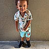 child, boy, smiling, curly_hair, standing, mickey_mouse, shorts, shirt, sneakers, outdoor, wall, concrete, texture, shadow, playful, cute, portrait, sunlight, happy, casual