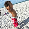child, beach, sand, ocean, sky, clouds, sunlight, curly_hair, swim_shorts, red_clothing, standing, barefoot, summer, outdoor, water_bottle, footprints, vacation, playful, sunny, relaxation