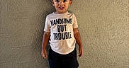 Azyaire is registered to the contest to win money with this photo: boy, casual_clothing, child, concrete_floor, curly_hair, expression, footwear, nike, outdoor, person, playful, portrait, shirt, shoes, shorts, standing, text_on_clothing, toddler, wall, young_child