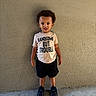 toddler, child, boy, standing, shirt, shorts, shoes, nike, text_on_clothing, wall, concrete_floor, curly_hair, expression, casual_clothing, outdoor, portrait, young_child, person, footwear, playful