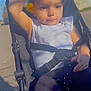 toddler, stroller, child, outdoor, sunlight, flower, yellow_flower, waving, white_shirt, black_pants, seatbelt, curly_hair, blue_sky, stone_wall, sidewalk, shadow, daytime, person, baby, casual_clothing